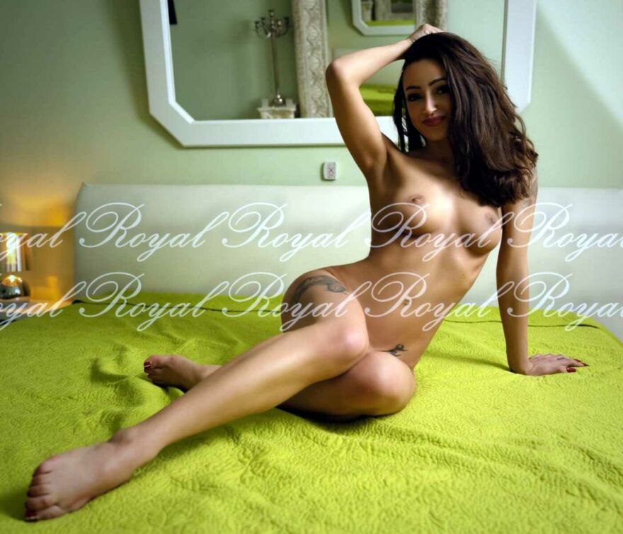 Die Hure Stella aus dem Bordell Royal sitzt anmutig und völlig nackt auf einem Bett und schaut lächelnd in die Kamera