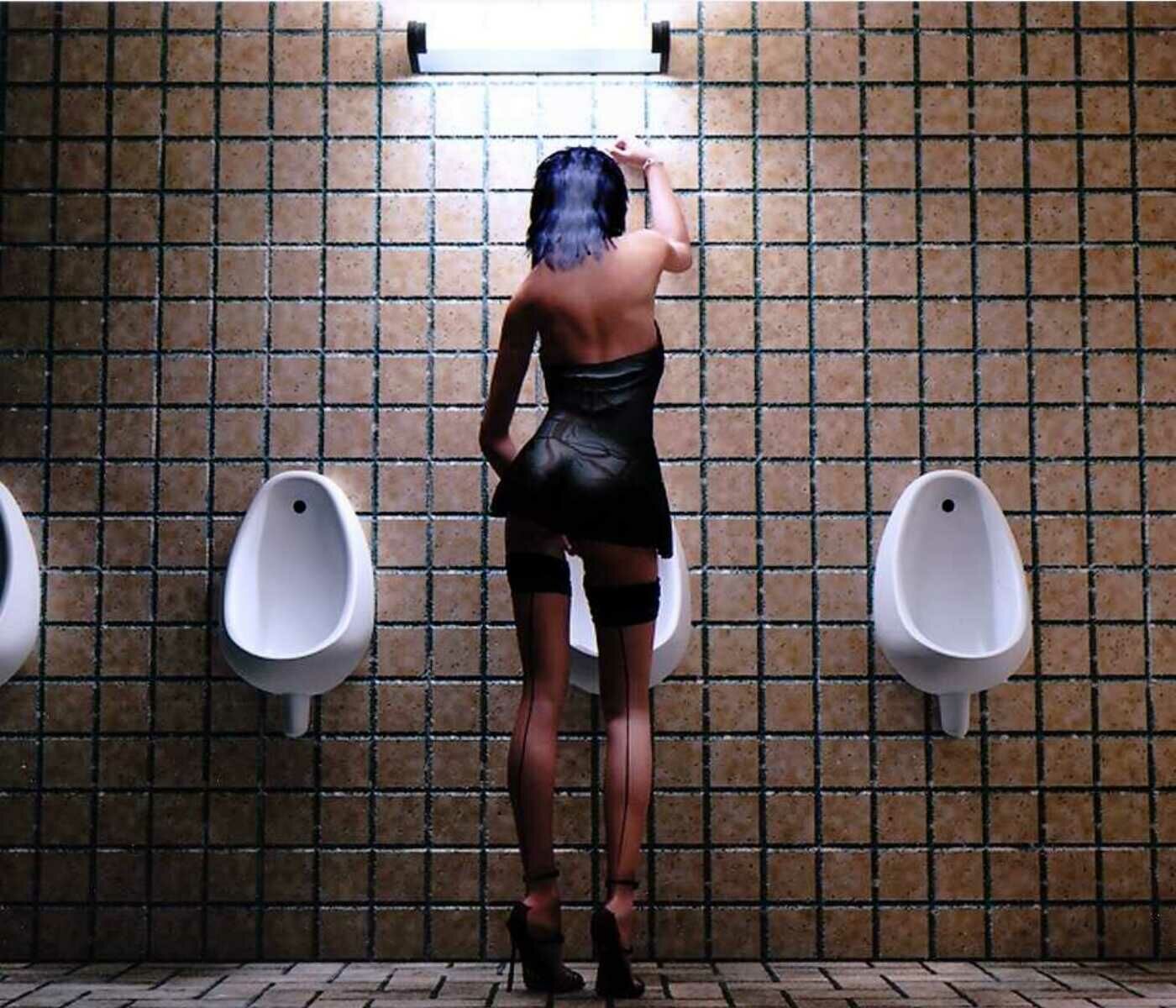 Ein transsexueller Mensch in Strapsen steht vor einem Pissoir