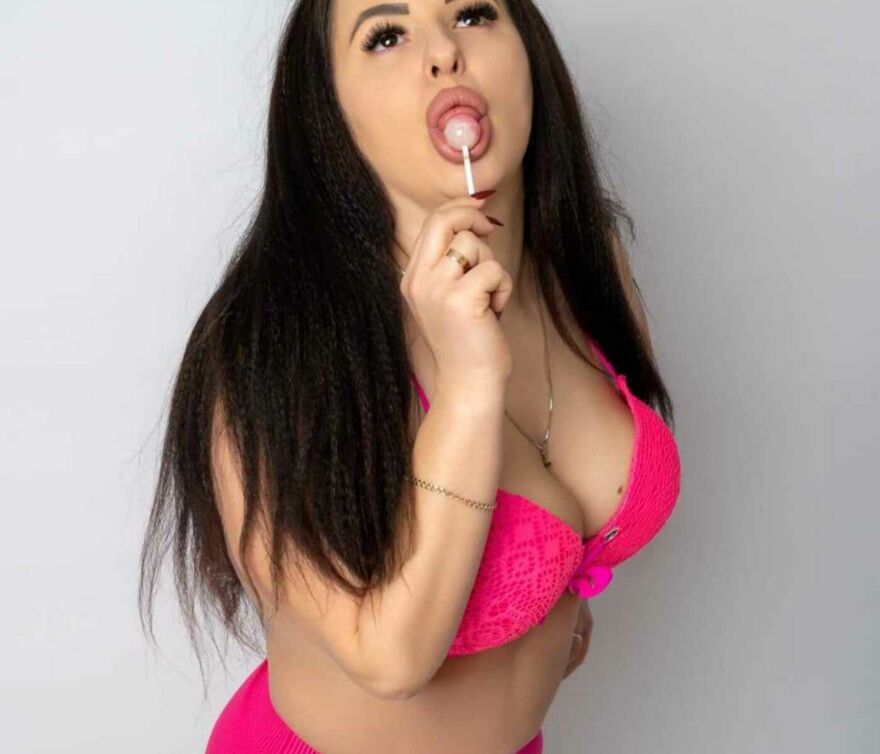 Das vollbusige Escort-Girl Natascha steht vor einer Mauer und lutscht an einem Lolli